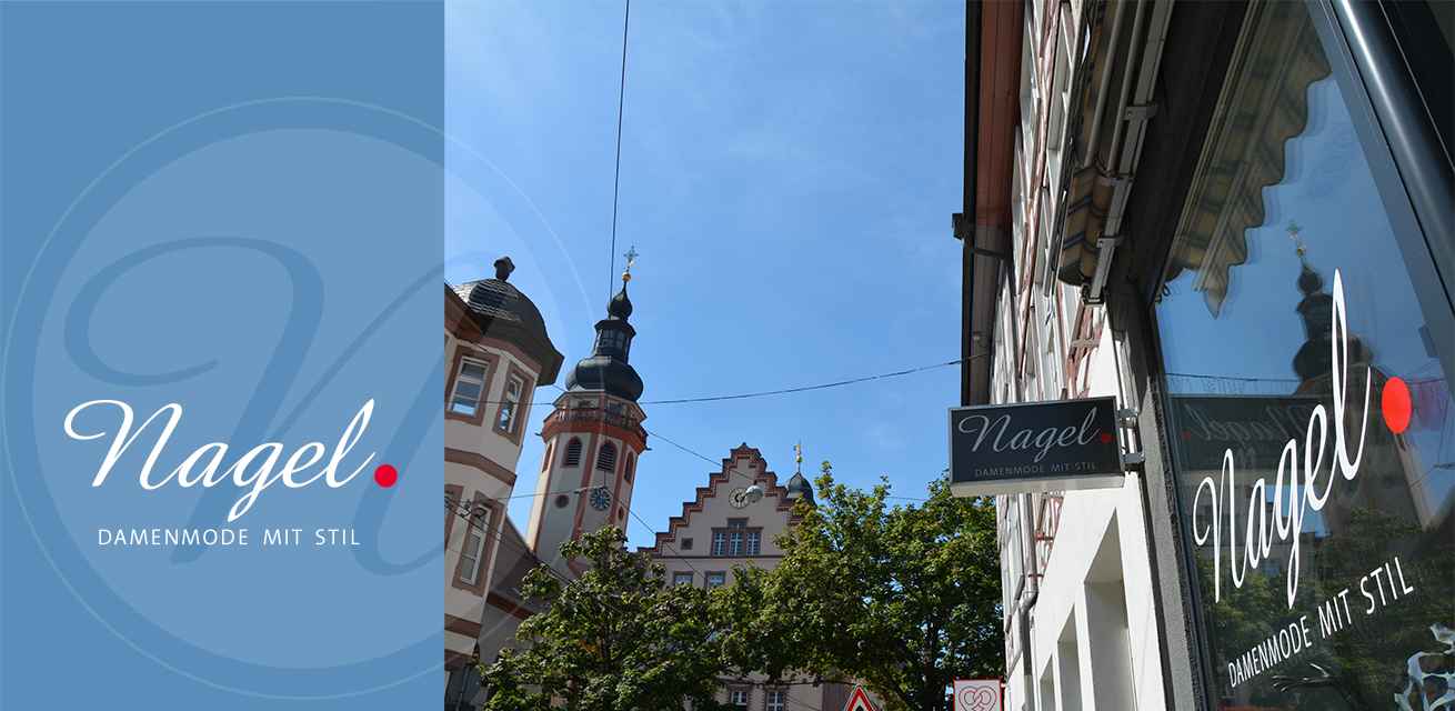 Ein Geschäftsschild mit der Aufschrift "Nagel. Damenmode mit Stil" hängt an einem Gebäude. Im Hintergrund sind ein Kirchturm und historische Fachwerkhäuser bei klarem Himmel sichtbar.