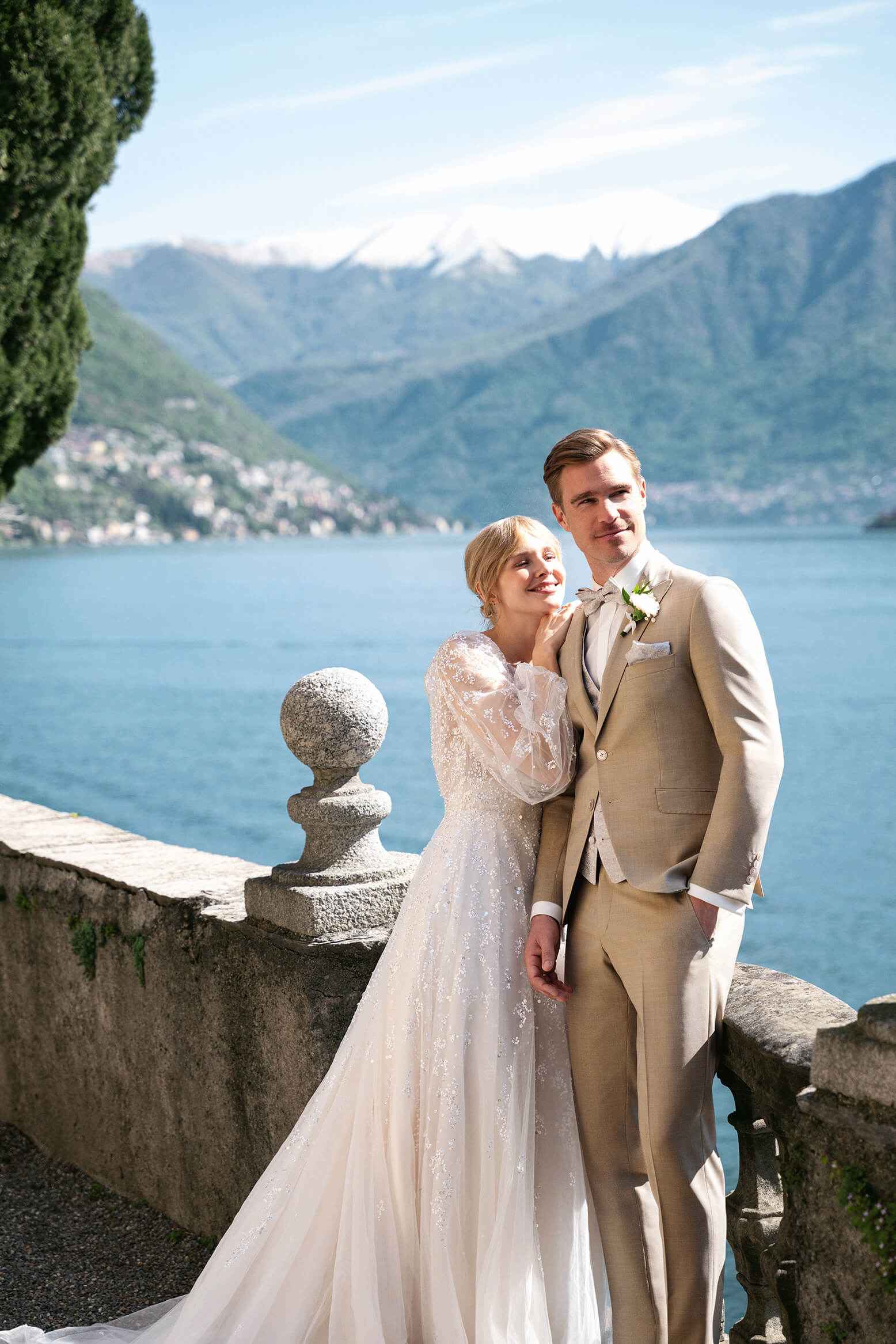 Brautpaar posiert elegant an einer Steinbalustrade mit Blick auf einen See und von Bergen umgeben. Der Bräutigam trägt einen beigen Anzug, die Braut ein weißes Kleid.