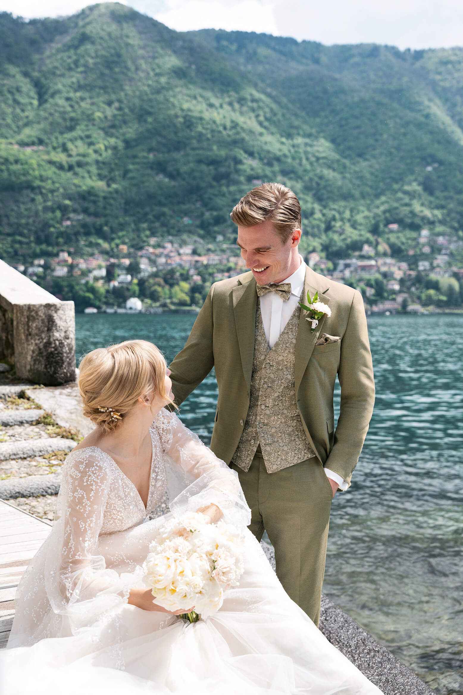 Brautpaar lächelt am Seeufer, Frau im Brautkleid mit Blumen, Mann im Anzug, umgeben von Bergen und Wasser, sonniger Tag.