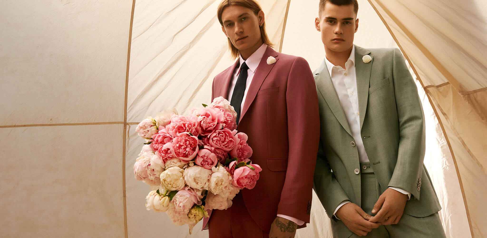 Zwei Männer in eleganten Anzügen stehen nebeneinander; der linke hält einen großen Strauß rosa und weißer Blumen. Der Hintergrund ist ein helles Zeltinnere.