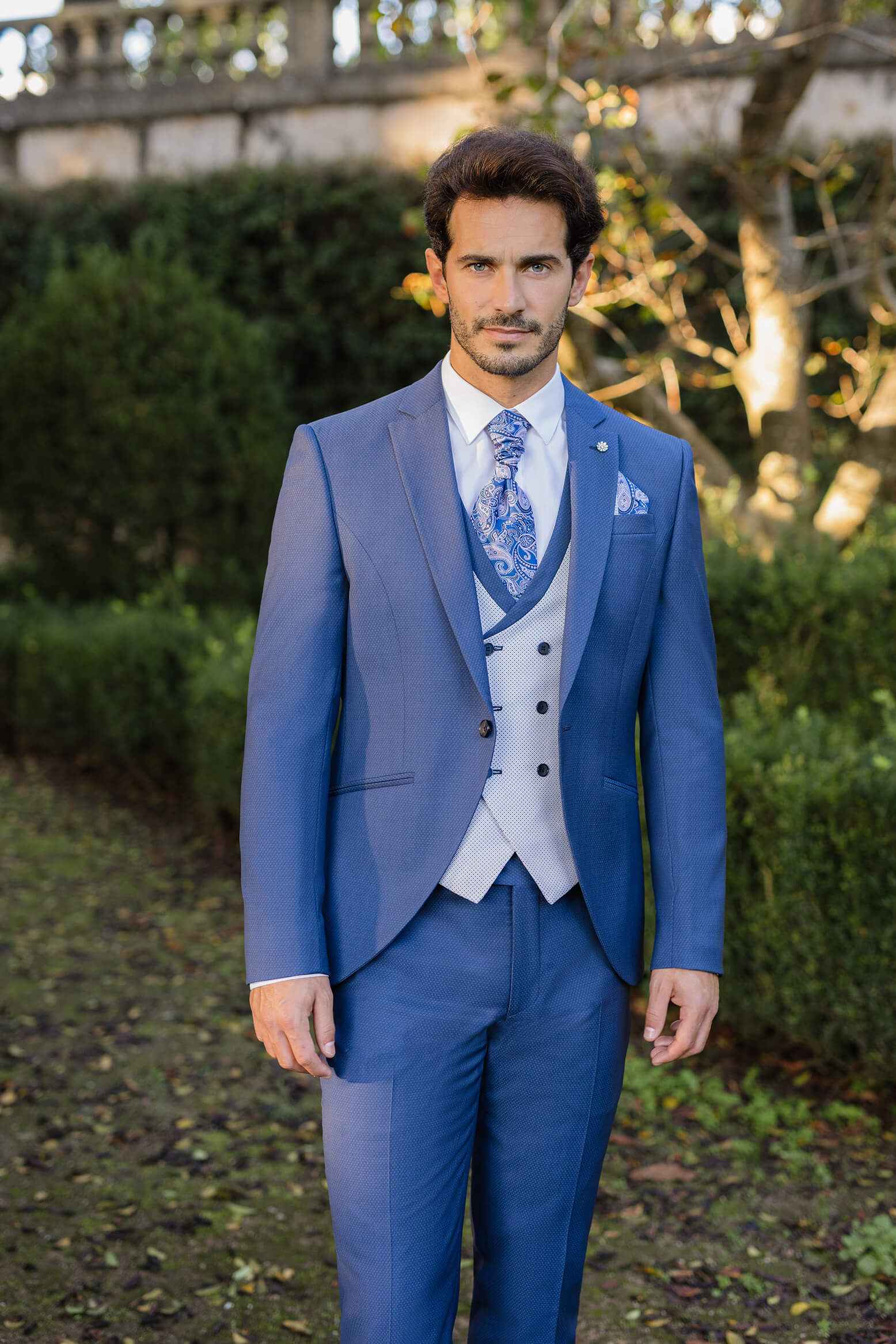 Ein Mann trägt einen eleganten blauen Anzug mit Weste und gemusterter Krawatte, steht im Freien vor einer grünen Hecke und einem Baum.
