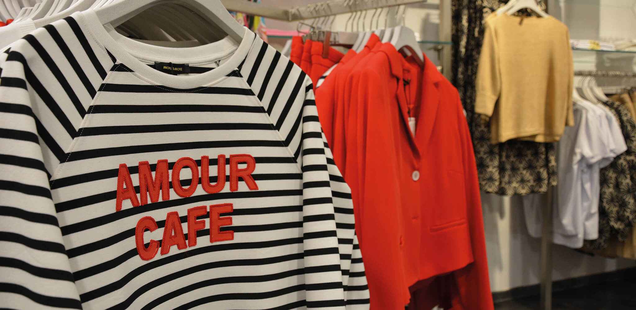 Gestreifter Pullover mit rotem Schriftzug "AMOUR CAFE" hängt auf einem Kleiderbügel. Im Hintergrund hängen rote, beige und gemusterte Bekleidungsstücke in einem Geschäft.