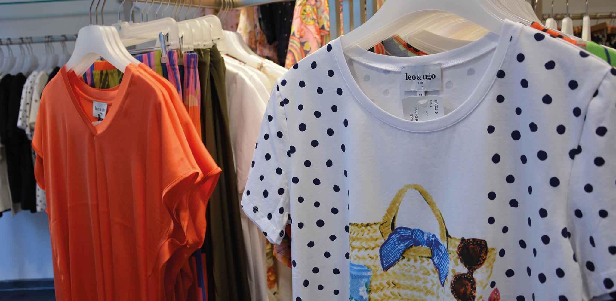 Ein weißes T-Shirt mit blauen Punkten und Taschenaufdruck hängt neben einem orangefarbenen Shirt. Im Hintergrund hängen weitere bunte Kleidungsstücke. Text auf dem Etikett: "leo & ugo". Preis: €79,99.