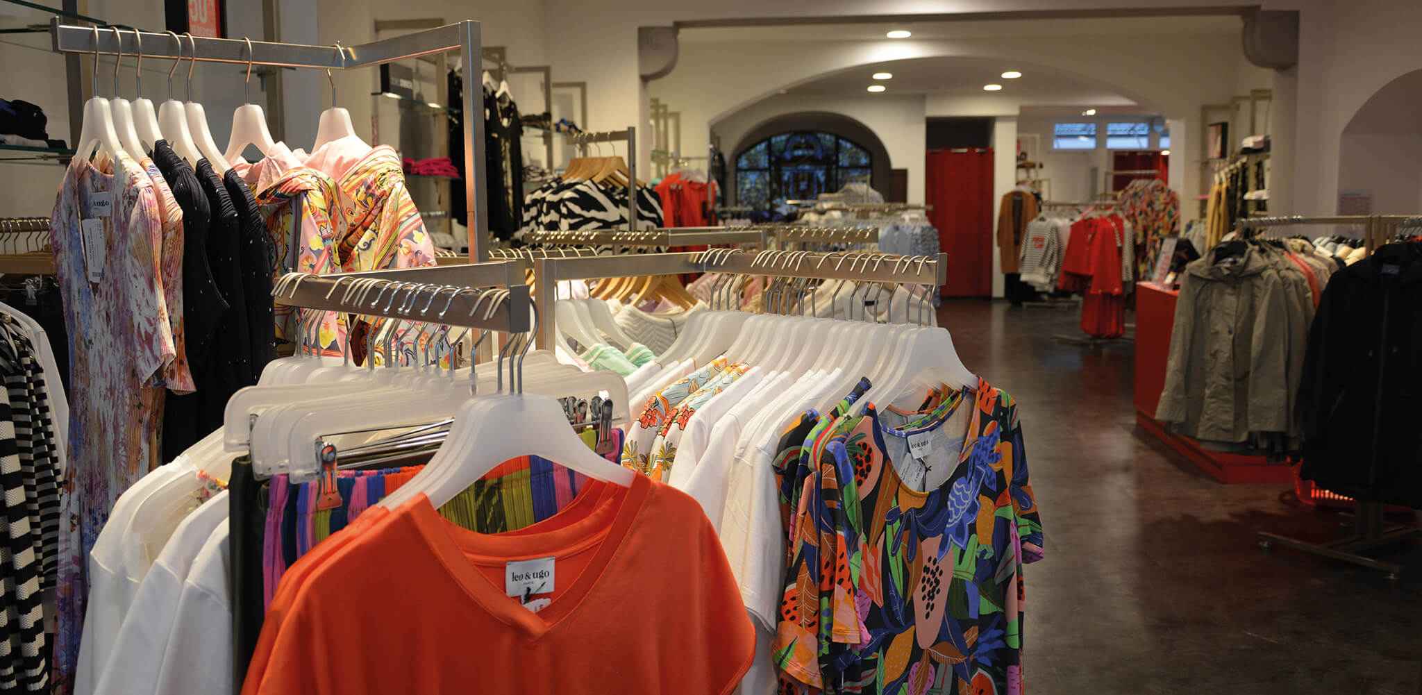 Kleidung hängt auf Kleiderbügeln in einem Geschäft. Verschiedene bunte Shirts und Muster sind zu sehen. Der Laden hat eine moderne Einrichtung und ist gut beleuchtet.