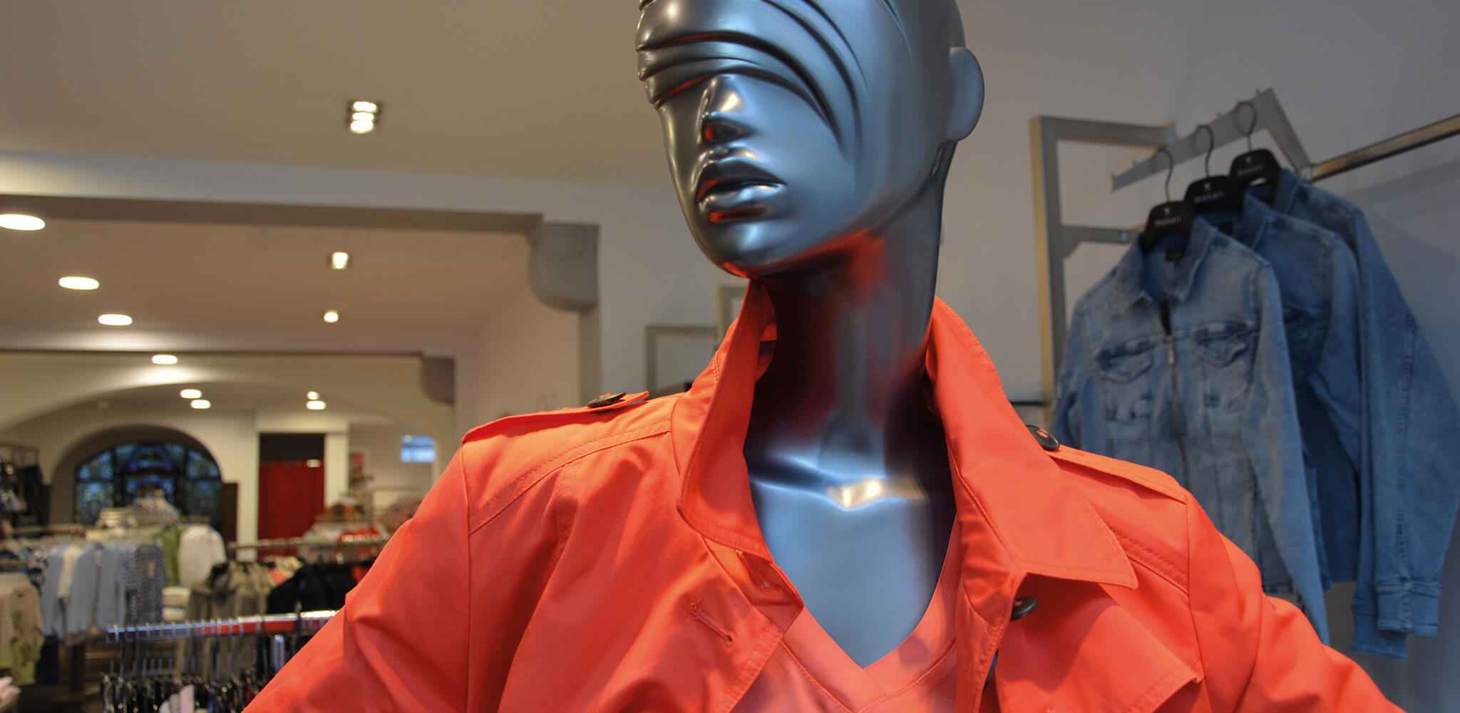Schaufensterpuppe trägt einen leuchtend orangefarbenen Mantel, steht still in einem beleuchteten Bekleidungsgeschäft mit Jeansjacken im Hintergrund und zeigt dessen Modekollektionen.