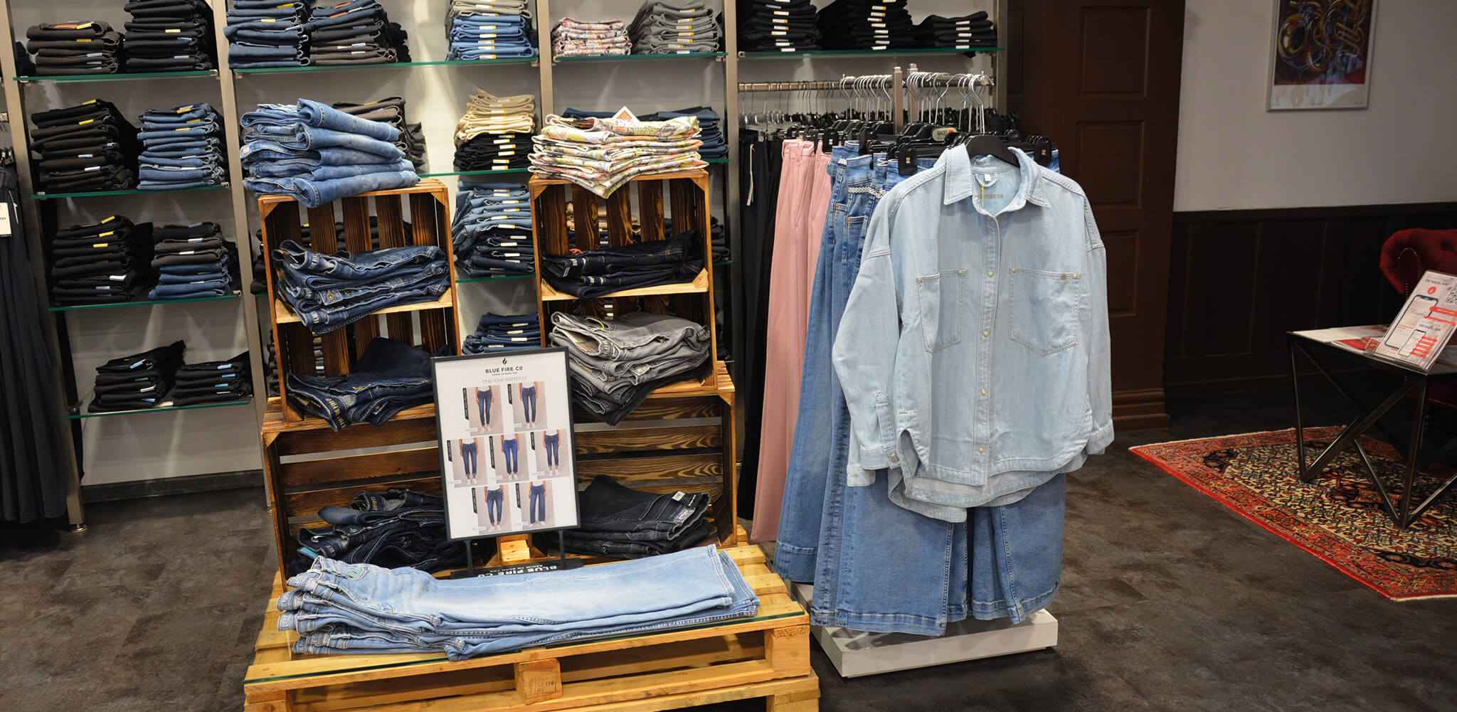 Jeanshosen liegen gefaltet auf Holzregalen in einem Bekleidungsgeschäft. Daneben hängt ein Jeanshemd auf einem Ständer. Ein Teppich liegt auf dem dunklen Boden. Text: „Blue Fire Co.“