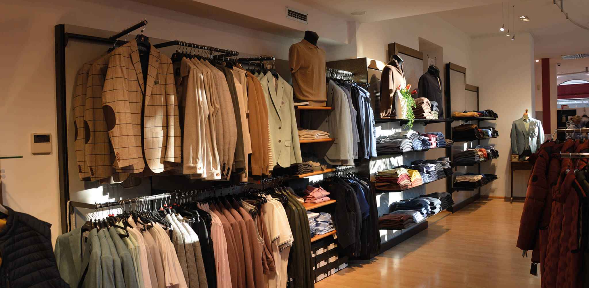 Jacketts und Hemden hängen ordentlich auf Kleiderständern. Dahinter stapeln sich Sweatshirts und Pullover. Die Umgebung ist ein gut beleuchtetes Bekleidungsgeschäft mit Holzboden.