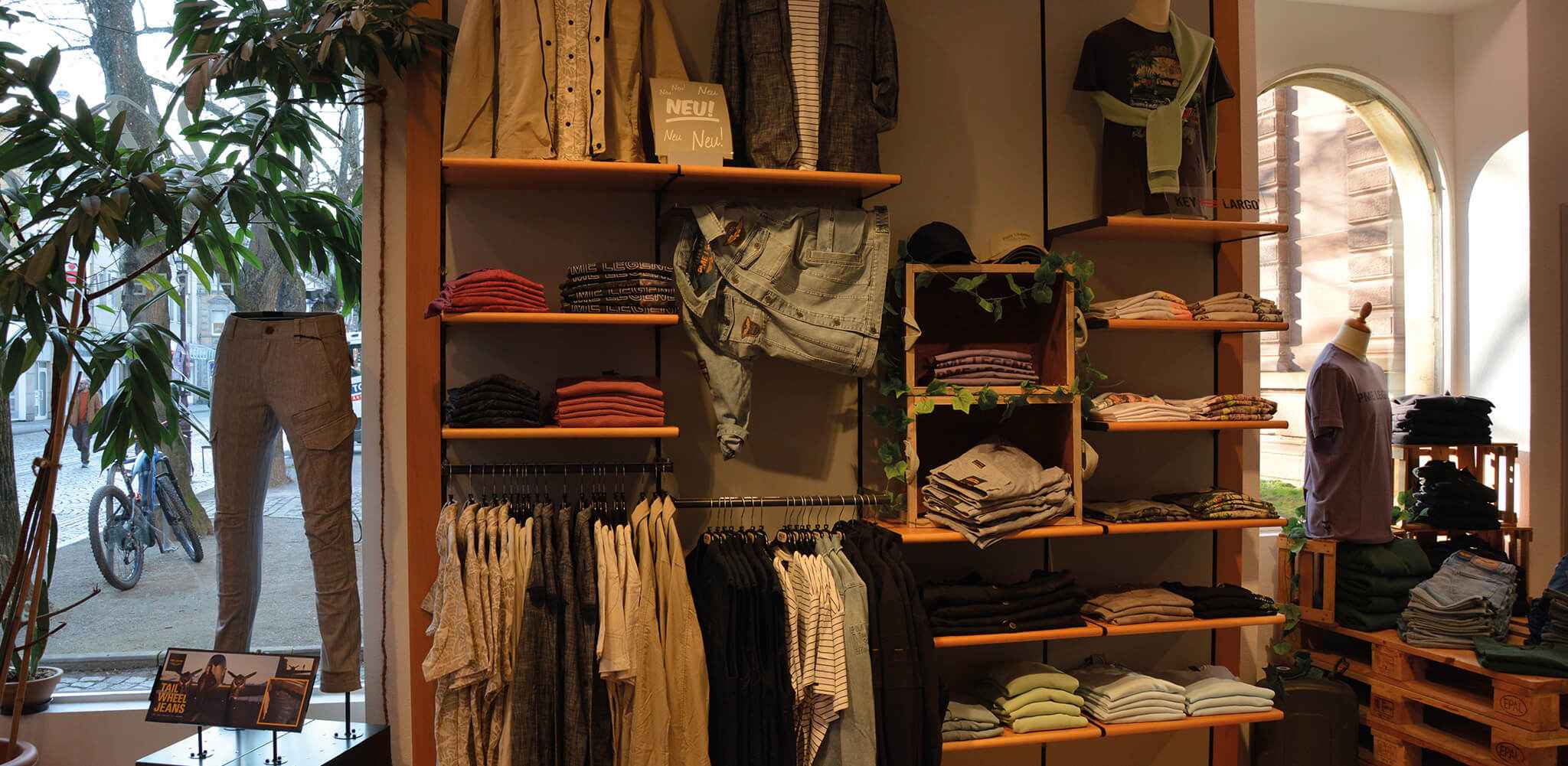 Kleidung wird auf Regalen und Mannequins präsentiert, umgeben von Pflanzen in einem Geschäft. Text: "NEU! Neu Neu!" und "TAIL WHEEL JEANS". Draußen sind Bäume und ein Fahrrad sichtbar.