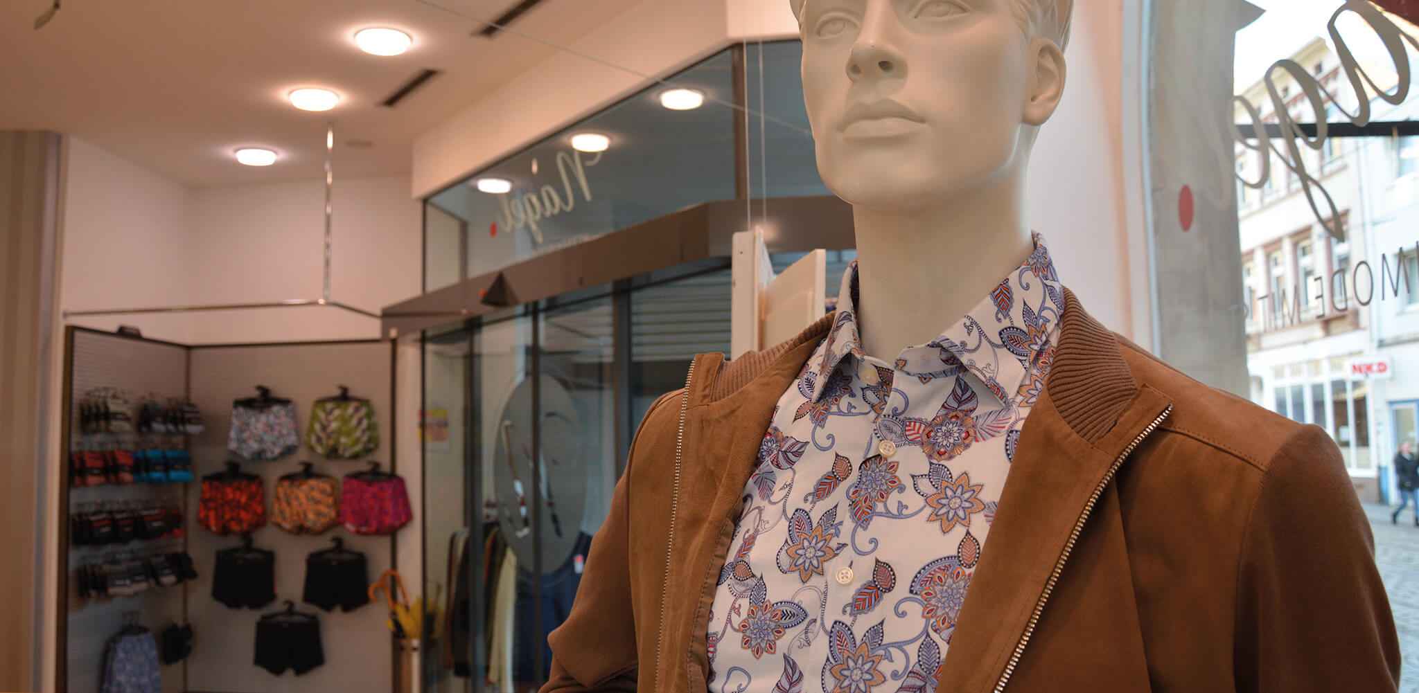 Schaufensterpuppe trägt gemustertes Hemd und braune Jacke, steht in bekleideter Einkaufspassage. Im Hintergrund bunte Shorts und Hemden, Schaufenster zeigt Worte: "MODEMIT" und "Nego".