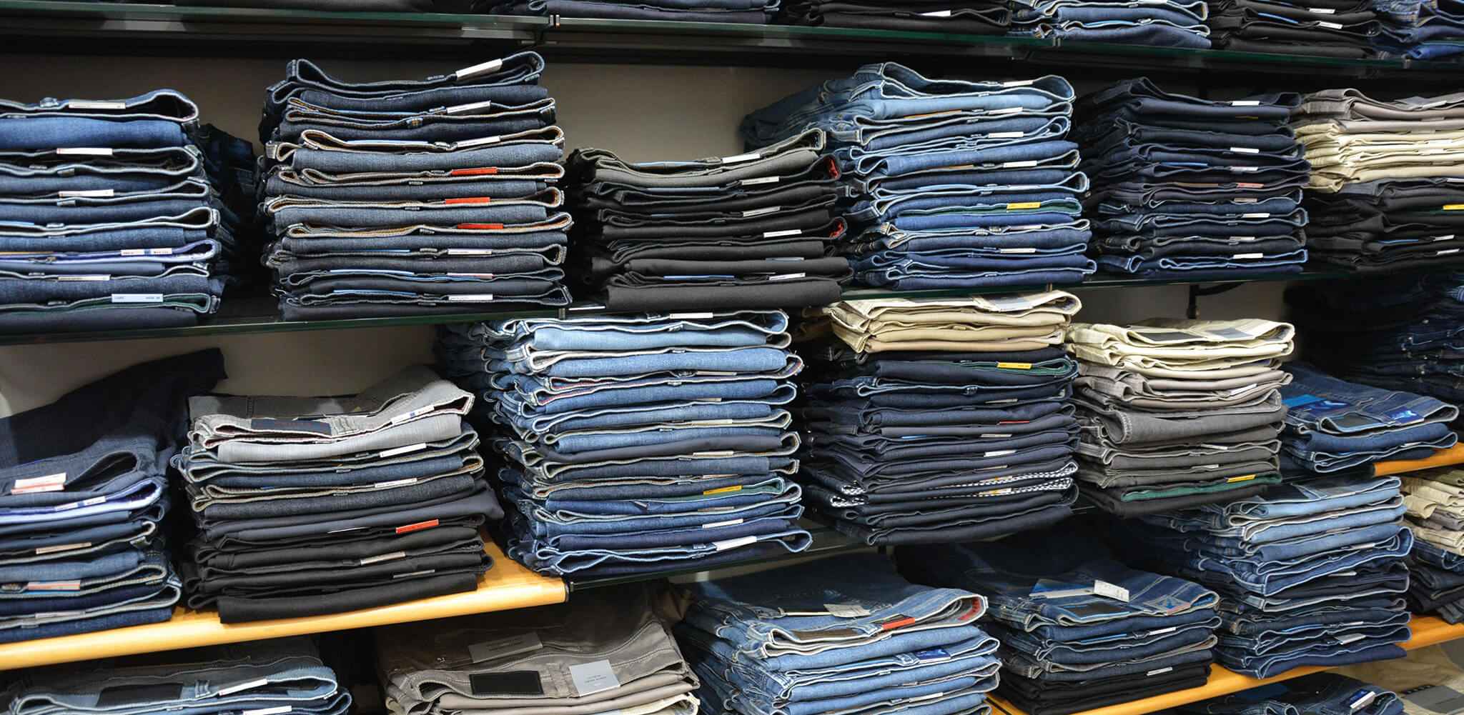 Mehrere Stapel gefalteter Jeans liegen ordentlich auf Regalen in einem Laden, umgeben von anderen Kleidungsstücken und verschiedenen Farbtönen, hauptsächlich Blau und Schwarz.