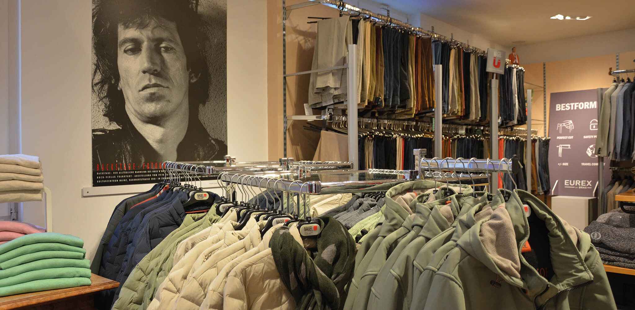 Kleider auf Kleiderständer hängen in einem Kleidungsgeschäft. Im Hintergrund sind Hosen auf Regalen. Ein großes Poster zeigt ein Schwarz-Weiß-Foto eines Mannes. Text: "BACKSTAGE A" und "BESTFORM."