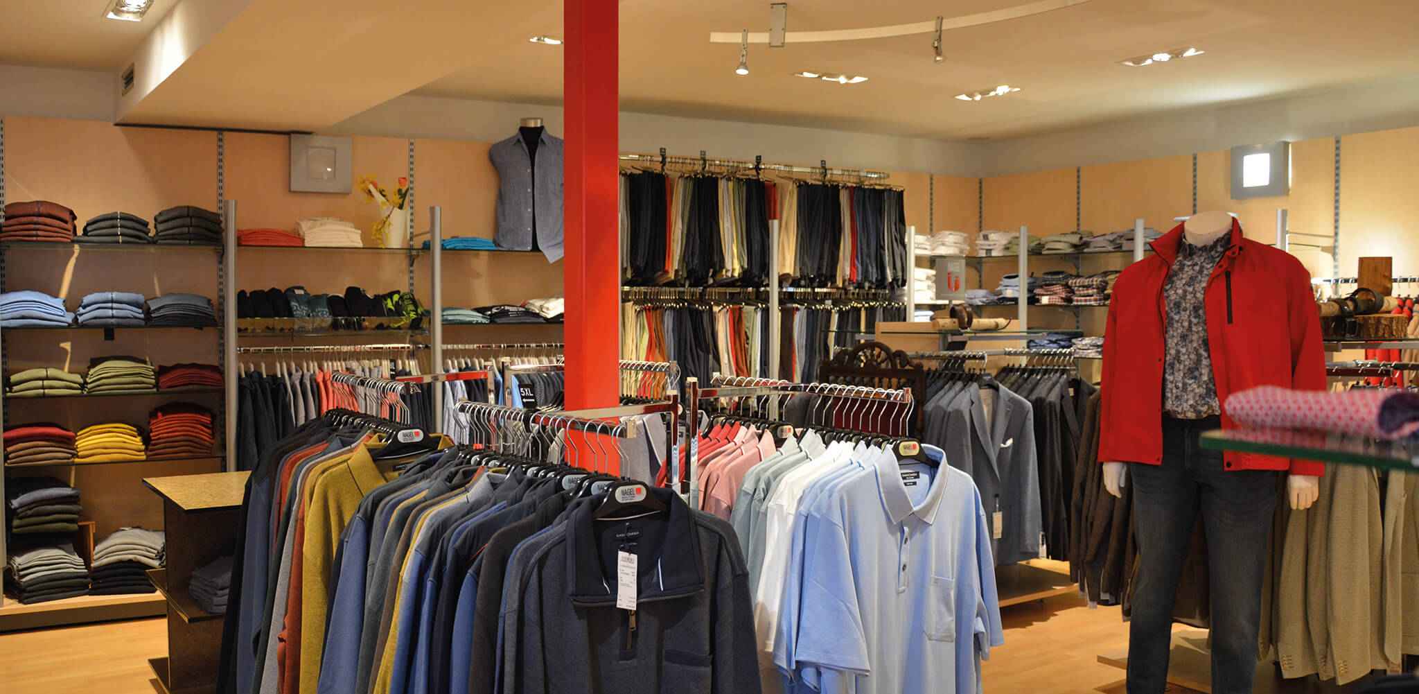 Kleider hängen und liegen in einem hell beleuchteten Kleidungsgeschäft; ein Mannequin trägt eine rote Jacke, umgeben von Hemden, Hosen und Pullovern in Regalen.