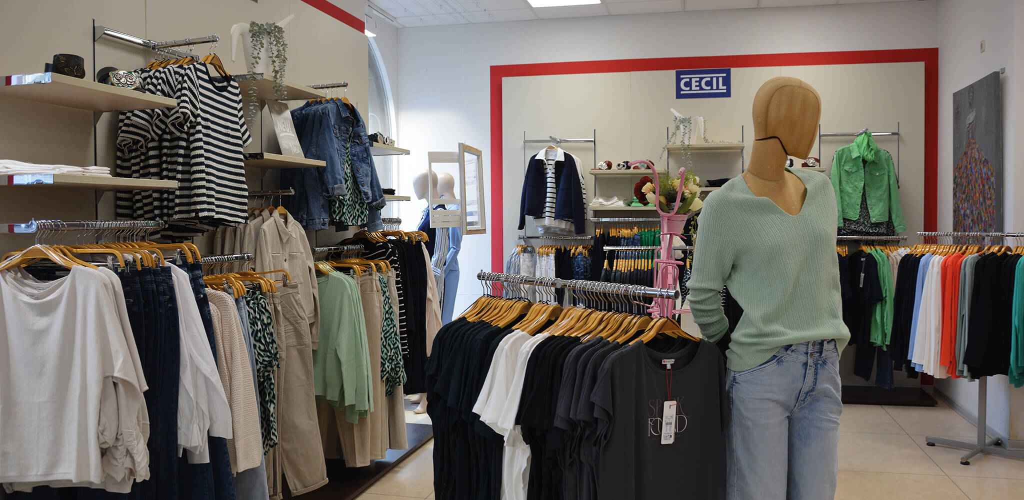 Eine Schaufensterpuppe trägt einen hellgrünen Pullover und Jeans, umgeben von ordentlich hängender Kleidung in einem Geschäft. Oben an der Wand steht "CECIL".