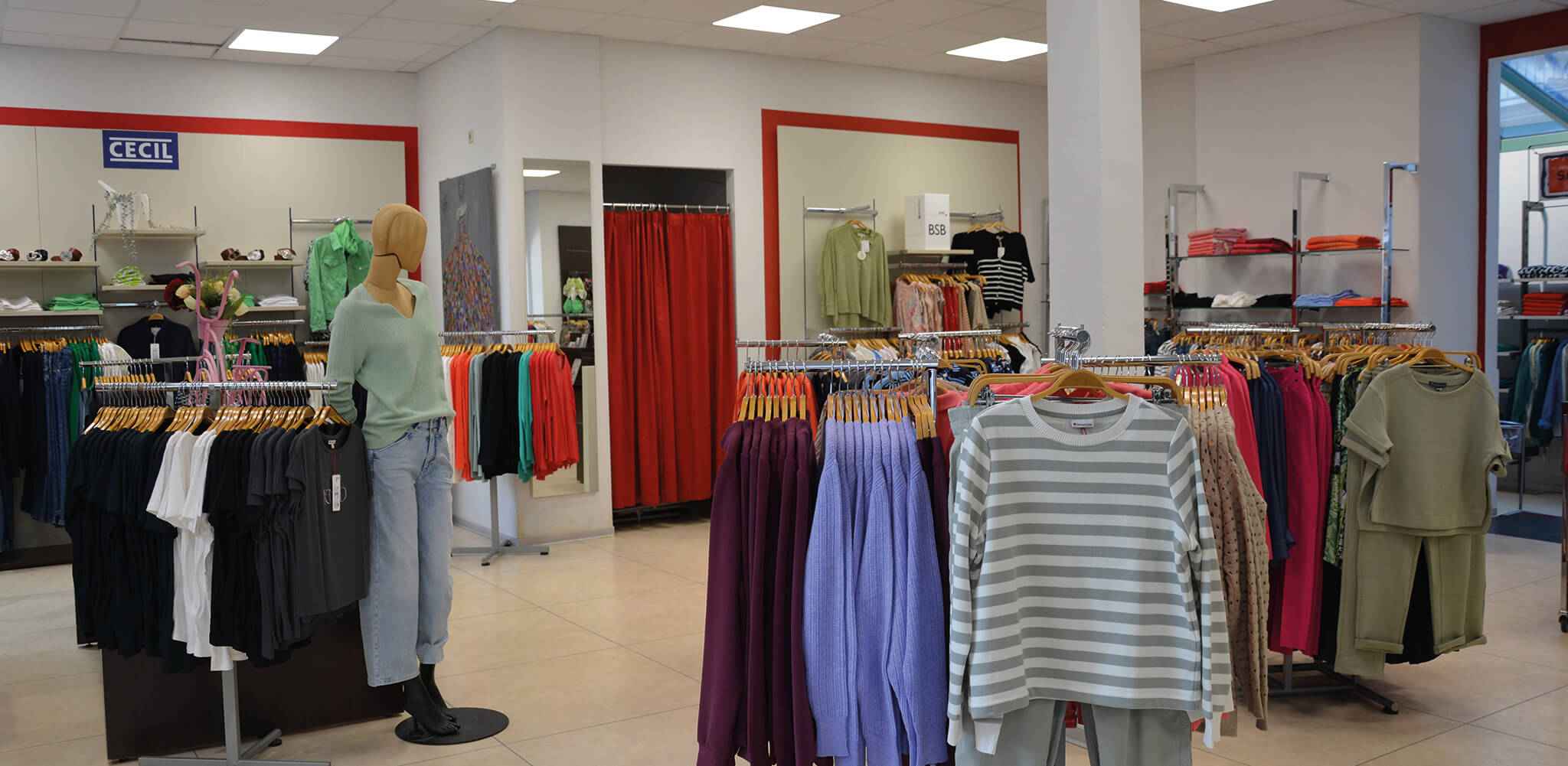 Eine Schaufensterpuppe steht in einem Bekleidungsgeschäft, umgeben von Kleiderständern mit bunter Kleidung. Im Hintergrund ist das Schild „CECiL“ sichtbar und rote Vorhänge.