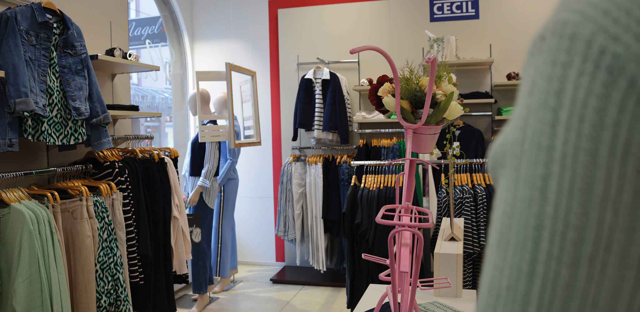 Kleider hängen ordentlich auf Holzkleiderbügeln, ein pinker Ständer mit Blumen im Vordergrund, innerhalb eines gut beleuchteten CECIL Bekleidungsgeschäfts.