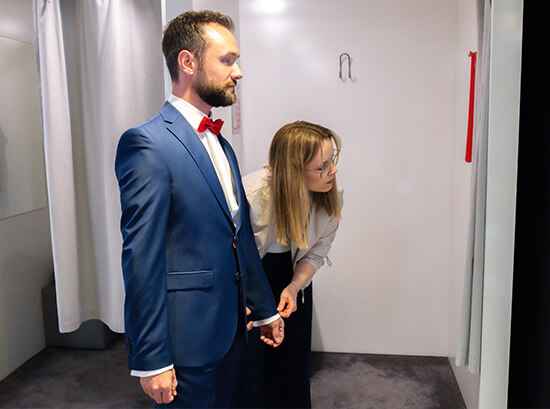 Ein Mann im blauen Anzug steht, während eine Frau seinen Ärmel anpasst; sie befinden sich in einem Umkleideraum mit grauem Boden und weißen Wänden.