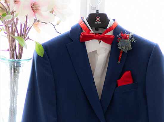 Ein blauer Anzug mit roter Fliege und Einstecktuch hängt an einem Kleiderbügel, geschmückt mit Boutonniere. Im Hintergrund steht eine Vase mit rosa Blumen.