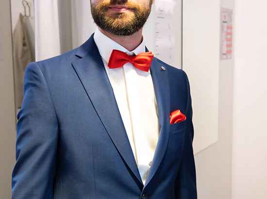 Ein Mann trägt einen dunkelblauen Anzug mit roter Fliege und Einstecktuch, steht in einem modernen Raum mit weißen Wänden und einem aufgehängten Kleidungsstück.