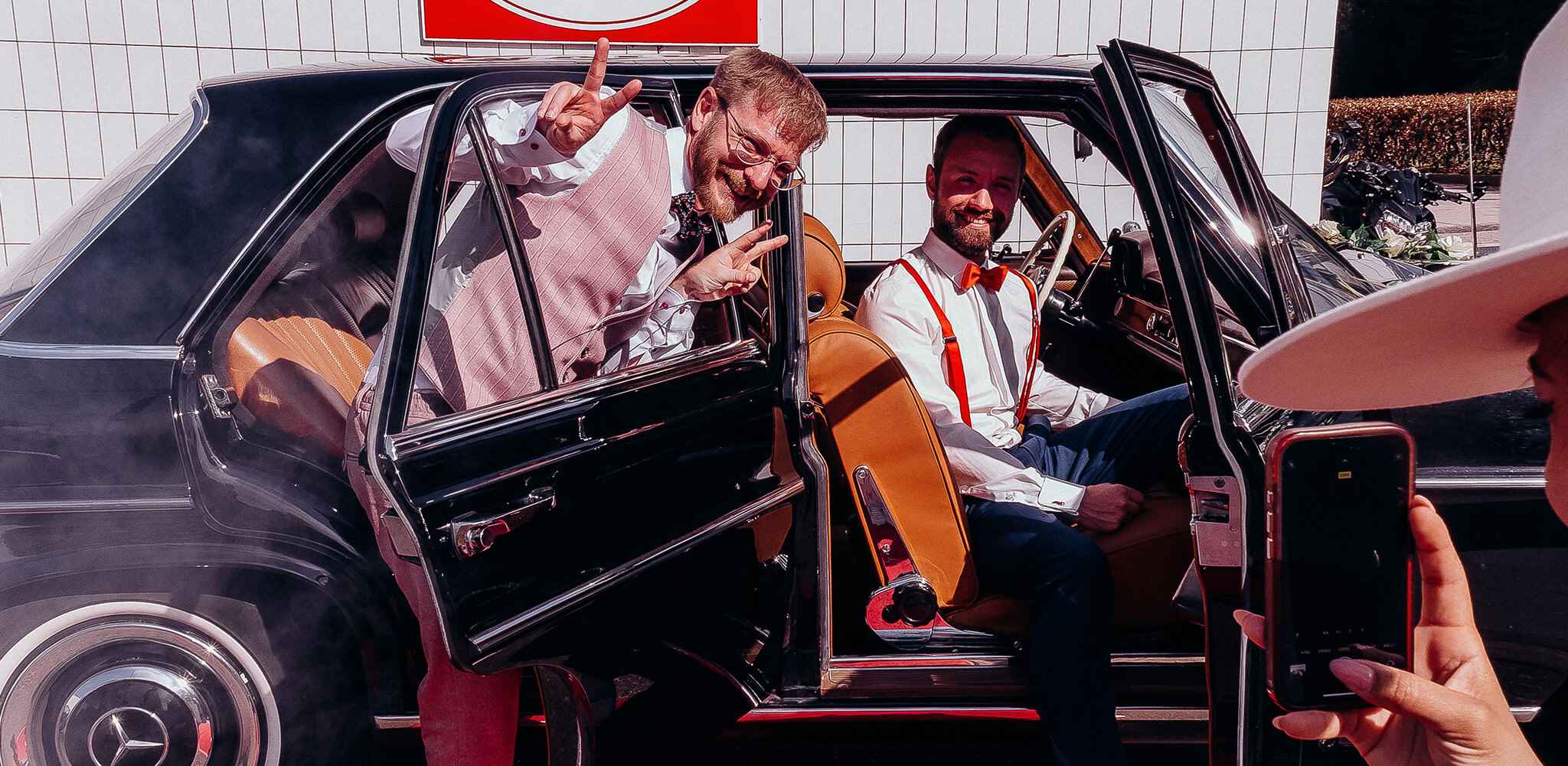 Ein schwarzes Auto steht mit geöffneter Tür vor einer weißen Wand, während zwei Männer in eleganter Kleidung posieren. Eine Person fotografiert sie mit einem Smartphone.