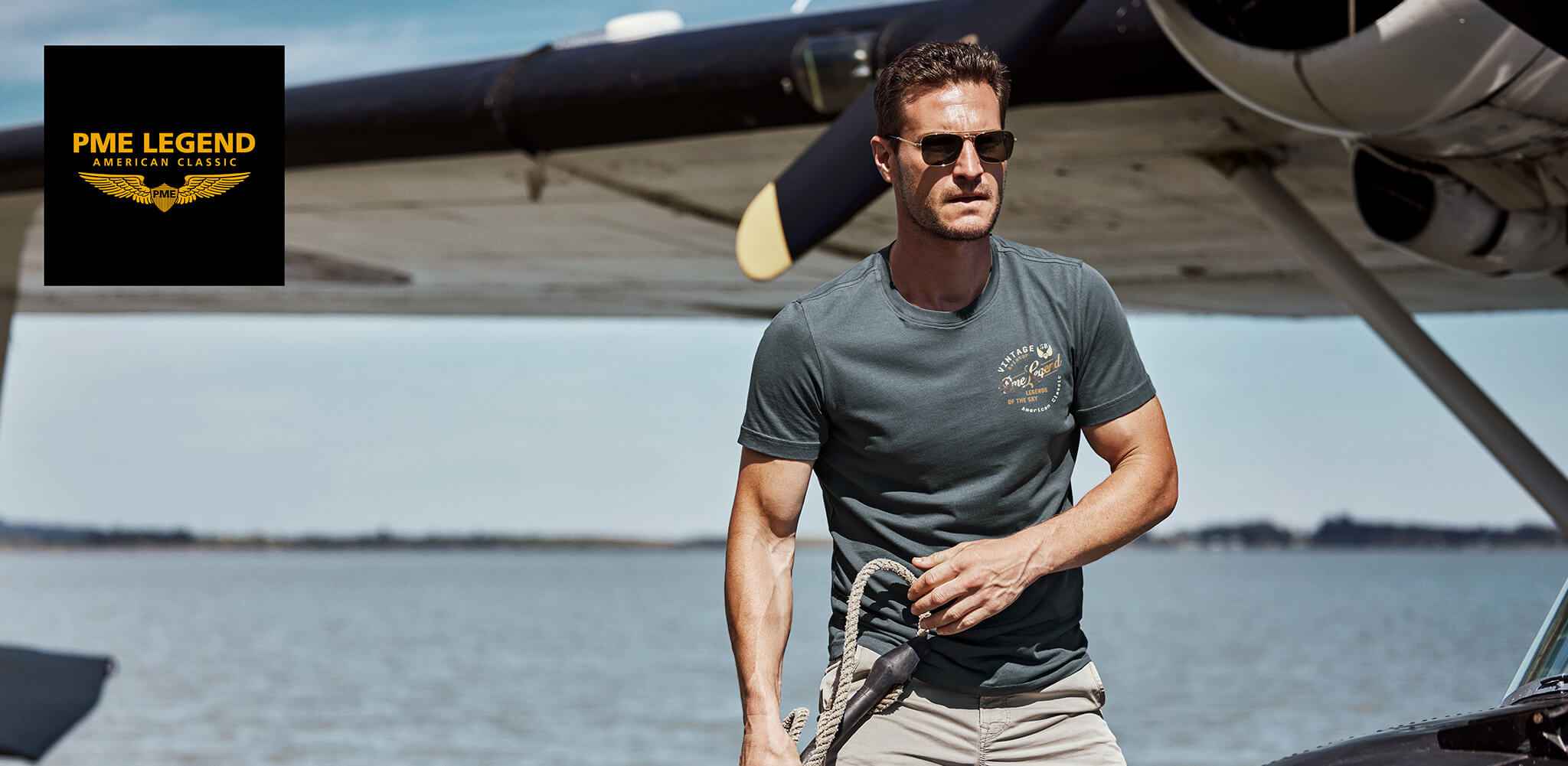 Ein Mann hält ein Seil, während er vor einem Wasserflugzeug am Ufer steht. Text: „PME LEGEND AMERICAN CLASSIC“ und auf dem T-Shirt: „VINTAGE PME LEGEND THE SKY IS OUR HOME“.