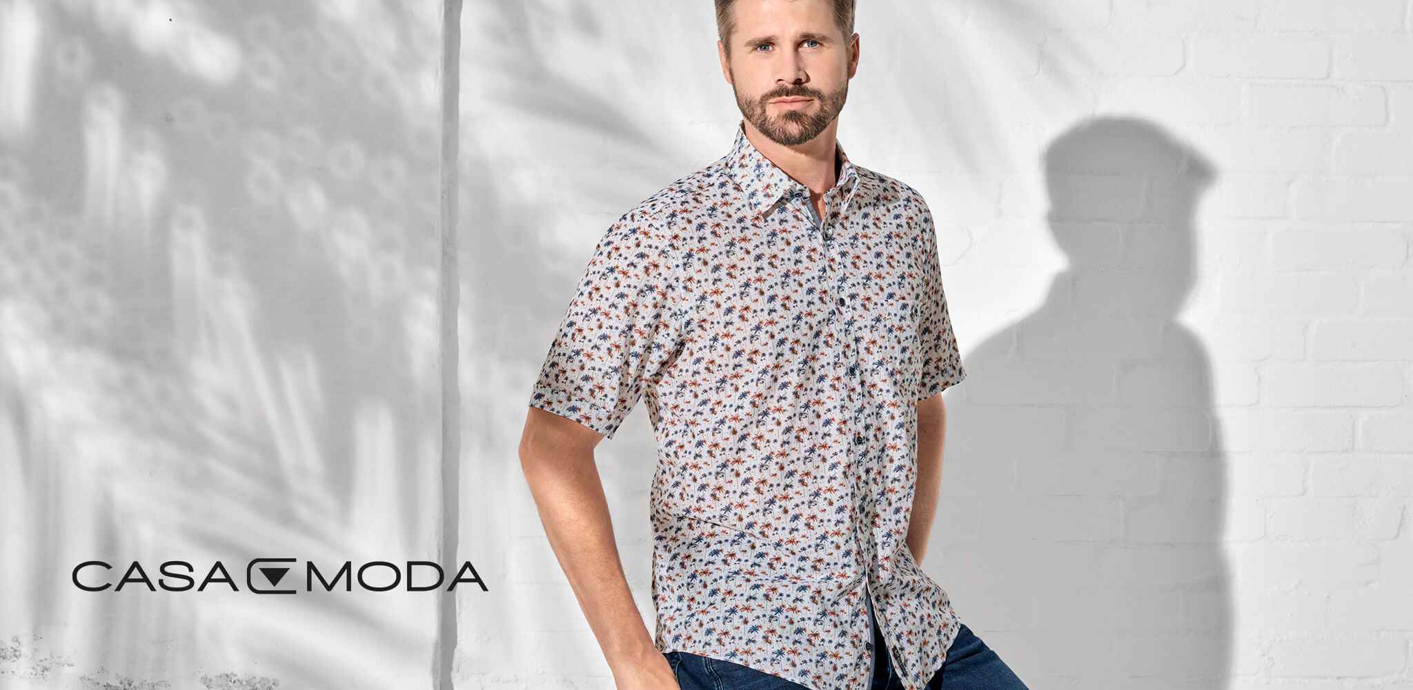 Ein Mann trägt ein gemustertes Hemd, steht vor einer weißen Wand. Schatten und Licht erzeugen Muster. Text: "CASA MODA" links unten.